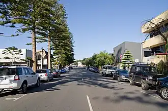 Street with parked cars and trees on both sides, surrounded by small buildings