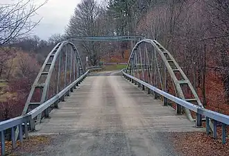 A metal bridge with two arched sides, forking gently over the course of their arch, braced at the top by two metal bars, and a wooden deck, in a semi-wooded area at a time of year when no trees are in leaf and dead leaves cover the ground.