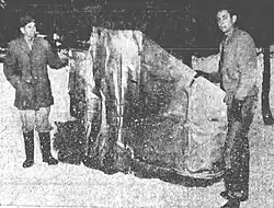Two men holding up a crumpled sheet of metal on snow-covered ground