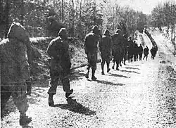 A line of soldiers walk down a gravel road into the distance