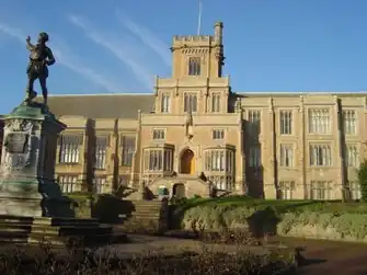 Photograph of a Gothic Revival building. The walls are light brown and the pitched, grey-slate roof has algae growing on it. There is a wide chimney on each side. There are five large multi-segment windows on each story on the left section and on the right section. Both sections have two and three stories and are separated by a protruding central section, which has a total of five stories and a two-storey bay window on each side of an arched, wooden door, which is elevated. The door is above an outdoor double staircase, which has a simpler wooden door in the front. Above each bay window is a smaller window. Above the smaller windows is a narrower section with three large multi-segment windows, surmouned by a yet narrower section, forming a square tower and the highest storey of the building. The tower is crenellated and has one large multi-segment window, a flagpole without a flag in the centre, and a crenellated stone structure projecting from the right corner that resembles a crow's nest in that it could be used as a lookout point. In front of the building is a lawn that separates an embankment from a planted area. Steps lead down from the central part of the building to a war memorial in the form of a bronze statue of a uniformed, First World War British soldier looking backwards and pointing up towards the left, standing atop a white stone plinth with a relief of the city's heraldic achievement, an inscription on a plaque, and a relief of the lozenge of Dame Agnes Mellers. The war memorial is elevated with stone steps on all four sides, to separate an outer circle of shrubbery. Below the memorial is a paved stone floor.