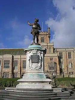 Photograph of a Gothic Revival building. The walls are light brown and the pitched roof, which has algae growing on it, is grey slate. There is a wide chimney on each side of the building. There are five large multi-segment windows on each story on the left section, and five large multi-segment windows on each story on the right section. The left and right sections have two and three stories respectively and are separated by a protruding central section, which has a total of five stories. The central section has a two-storey bay window on each side of an arched, wooden door, which is elevated. The door is above an outdoor double staircase, which has a simpler wooden door in the front. Above each bay window is a smaller window. Above the smaller windows is a narrower section with three large multi-segment windows, above which is a yet narrower section, forming a square tower and the highest storey of the building. The tower is crenellated and has one large multi-segment window, a flagpole without a flag in the centre, and a crenellated stone structure projecting from the right corner that resembles a crow's nest in that it could be used as a lookout point. In front of the building is a lawn that separates an embankment from a planted area. Steps lead down from the central part of the building to a war memorial, which is in the form of a bronze statue of a First World War uniformed British soldier looking backwards and pointing up towards the left, standing atop a white stone plinth that has a relief of the heraldic achievement of the city of Nottingham, an inscription on a plaque, and a relief of the lozenge of Dame Agnes Mellers. The war memorial is elevated with stone steps on all four sides, which separate an outer circle of shrubbery. Below the war memorial is a paved stone floor.
