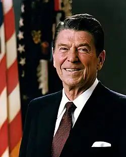 White House portrait of Reagan smiling in front of the U.S. and U.S. president flags, wearing a dark blue suit jacket with a white shirt and burgundy necktie.
