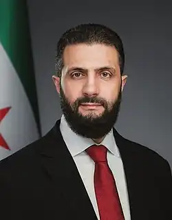 Headshot of al-Sharaa wearing a black suit jacket, a white shirt, and a necktie.