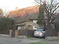 A wide stone cottage with a deep thatched roof illuminated by low sunlight. Two small rectangular windows can be seen on the right of the building, next to some trees and a car. A bush, fence and the roof obscure most of the rest of the cottage. There is a small chimney-stack on the left.