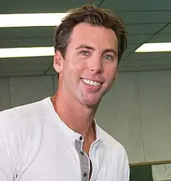 Profile of a smiling, brown haired young main, wearing sunglasses, wearing a yellow jacket tracksuit with a green collar, with a red medal ribbon around his neck.