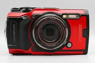 A red Olympus Tough TG-6 digital camera viewed from the front with a flash, and text indicating it is waterproof and shockproof.