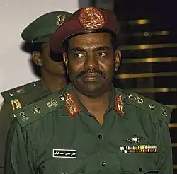 This is a coloured photograph of President Omar al-Bashir in 1990 in military attire, wearing a green uniform adorned with insignias, ribbons, and rank decorations. He is also wearing a maroon beret with an emblem at the centre. The individual has a composed yet stern expression, with a moustache and a focused gaze slightly off-camera. In the background, another person in a similar green uniform is partially visible, contributing to the formal and official atmosphere of the scene.