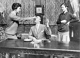 middle-aged white woman, standing, pulls the nose of a seated white man. A younger woman, standing, laughs