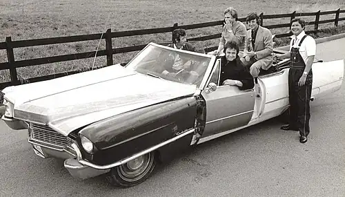 A group of four men sitting in a large car oddly constructed of mismatched parts. A fifth man in bib overalls is standing beside the car.