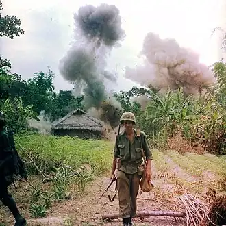 color photograph of a Marine calmly walking away from an exploding hut at the edge of a jungle