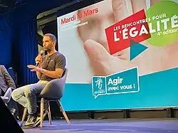 Ouissem is sitting on a chair on stage and speaking into a microphone; behind him is the poster of the event "Rencontres de l'Égalité"; He is wearing a black t-shirt and blue jeans