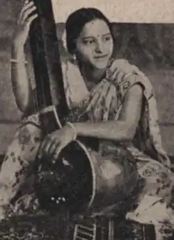 A young South Asian woman wearing a light-colored sari, seated, arms around a large stringed instrument