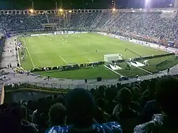 A football match taking place in a packed stadium with an athletics track, viewed from a stand behind one of the goals; it's night and the ground is floodlit.