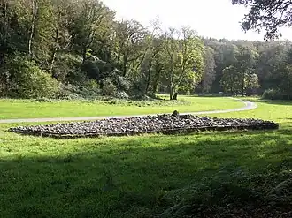 Rocks and boulders, forming a cairn, are retained by a short wall. Flat ground of short grass (the foreground in dappled shade and the rest in full sun) that surrounds the cairn is dissected by a pathway sweeping past and into the distance. In the background, trees, mainly in leaf, climb the steep sloped gorge.