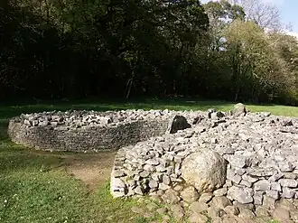 A short dry-stone wall retains boulders to form a cairn. The wall is missing at the front, right section, where the rubble has tumbled out, leaving a (previously covered) orthostat exposed. The wall forms a courtyard at the tumulus' entrance. Flat ground of short grass surrounds the cairn. The background is of shaded trees, mainly in leaf.