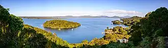 Paterson Inlet from up higher inland. The small, forested Iona Island is visible not far from land, and many small sailboats dot the water. Surrounding land is lush, with some houses visible through the trees, and a dock on shore.