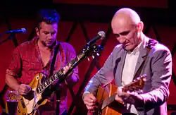 A man at left is playing an electric guitar, while 56-year-old Kelly plays his acoustic guitar. Both are looking down towards their own guitar, each uses a plectrum in their right hand while the left is on the fret board. In front of each is a microphone on its stand.