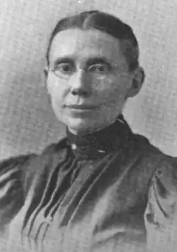 A white woman with dark hair parted center and dressed severely back to the nape; she is wearing glasses and a dark garment with a high collar, pleated bodice, and puffed sleeves