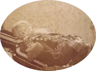 Photograph showing a white-bearded man, hands folded on stomach, dressed in a uniform with sash and chain of office and lying in state on cushions with a book under the cushion at the head