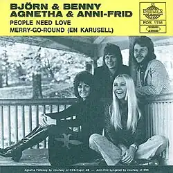 Two women are sitting at the front of the porch, while two men are standing at the back of the porch, both facing forward. The text with a yellow background reads "BJORN and BENNY, AGNETHA and ANNI-FRID. PEOPLE NEED LOVE, MERRY-GO-ROUND (EN KARUSELL)".