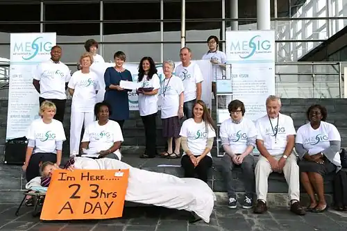 A group of 15 people posing in front of a building. One person lays on a cot holding a sign that reads, "I'm Here...23 hrs a day"