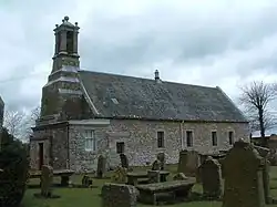 A grey stone chapel, with angular corners. An ornate bell tower is situated on the west of the building.