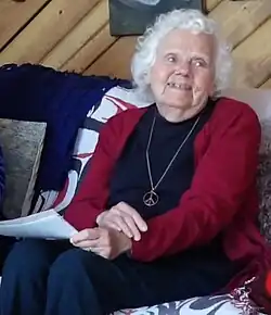 White-haired woman wearing a dark t-shirt under a red sweater and a long necklace with a peace sign