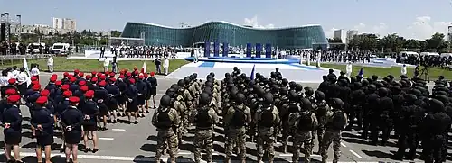A wide shot of a police ceremony outdoors. In the foreground, rows of officers in various uniforms (red berets, camouflage, and all-black tactical gear) stand in formation. In the background, a large, modern glass building and a stage with officials can be seen under a clear sky.