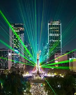 A stage with pyrotechnics and lights with a crowd dancing surrounded by skyscrapers.