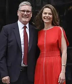 A woman in a red frock with her husband in a black suit standing in the front of a door and smiling.