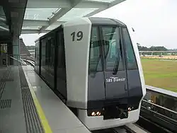 A white and dark blue Mitsubishi Crystal Mover train at the elevated platform of Punggol LRT station. The train, numbered 19, is stationary with its doors closed, and beyond the platform, an open grassy field and some construction or distant structures are visible.