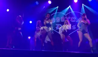 A picture of the Pussycat Dolls performing "React" during an appearance at GAY Nightclub in London. The stage is dimly lit with blue spotlights shining on each of the girls