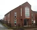 A modern building in two shades of red brick, behind a low black fence. White-framed three-paned windows line the side wall and flank the entrance—a double door sheltered below a protruding section supported on brick columns. This has a plaque reading "QUEEN STREET CHURCH CENTRE" and a pentagonal window.