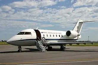 A twin-jet, high-tailplane passenger aircraft painted white above and grey below, parked on tarmac with its front stairs deployed