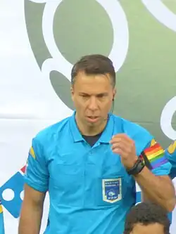 A man wearing a sky blue short sleeve shirt with a league patch on the left breast pocket and a rainbow armband on his left sleeve. He has a watch on his left wrist and he is raising that arm. He is wearing an earpiece on his left ear.