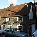 A small, two-storey flint cottage with a steep tiled roof, attached to another of the same height to the left. It has a white door and four irregularly spaced windows (edged in red brick), the upper two of which have window-boxes. A white gate is set into a flint wall attached to the right side of the cottage.