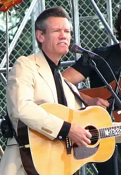 Randy Travis, wearing a tan suit and singing into a microphone while playing an acoustic guitar