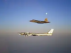 Aerial port view of two aircraft in flight, one on top of the other. The bottom aircraft is a four-engined propeller-driven aircraft, which is escorted by a jet fighter.
