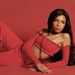 A photo of Lenae lying down and leaning on her elbow while looking off to the side. She is dressed in red; the floor and wall behind her are the same color.
