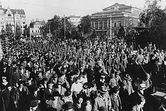 A crowd of civilians are standing in the foreground watching armed soldiers in the middle-ground marching alongside the street. Buildings of downtown Dresden can be seen in the background.