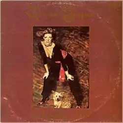 The artist's name is across the top in gold, styled writing over a maroon background. The title is styled similarly across the bottom, but all in lower case. The central image is the artist leading forward with her hands on her knees. A small dog is posed in front of her. She wears a black jump suit with a red scarf at her waist.