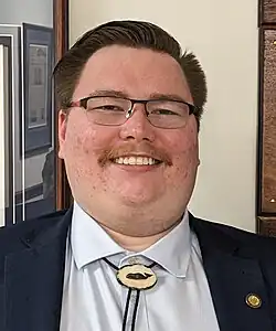 A man with a moustache, glasses, and a bolo tie smiling and looking directly at the camera