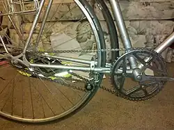 Close-up view of a retro-direct drivetrain on a bicycle. The chain loops over two freewheels of different sizes and is pulled taut by a tensioner mounted near the forward end of the chainstay.