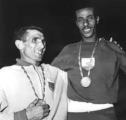 Abebe Bikila and Rhadi Ben Abdesselam standing with an arm around each other and their Olympic medals hanging from their necks. Both shown from about the waste up.