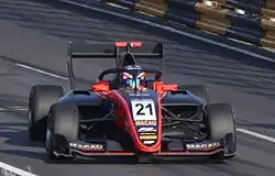 Front view of a black and orange racing car being driven at speed on a street circuit