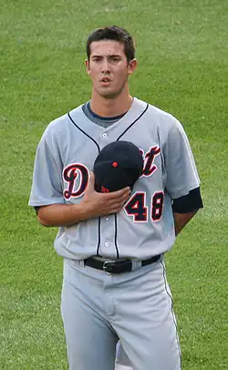 A man in a baseball uniform holds a baseball cap over his chest with his right hand.