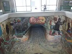 The mural features two hands holding water, which flows to other painted scenes. To the right, a man holding a pickaxe and another man giving water to a girl; to the left, a man offering water to an elderly woman and another operating construction equipment. On the floor, several aquatic species, including mollusks and plants, are painted