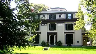 A white house with dark trim, including decorative elements. The house has three stories, and there is an octagonal element protruding above the top floor.