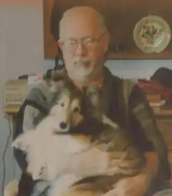 A balding elderly man with big glasses and a goatee holding a Shetland Sheepdog